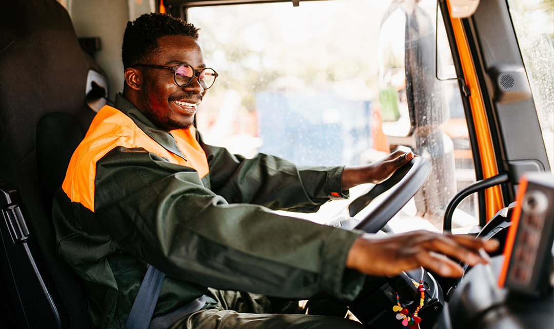 Conductor sentado en el asiento de piloto, con su mano izquierda sosteniendo el volante del vehículo y su mano derecha cerca de un panel de control, que indica el monitoreo de la flota, el consumo de combustible, la eficiencia del combustible y la seguridad de la flota.